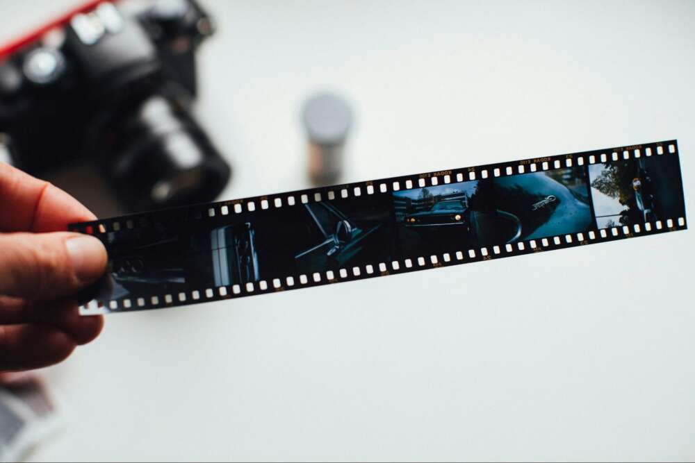A hand holding a strip of photographic film negatives with various images, with a blurred camera and film canister in the background on a white surface.