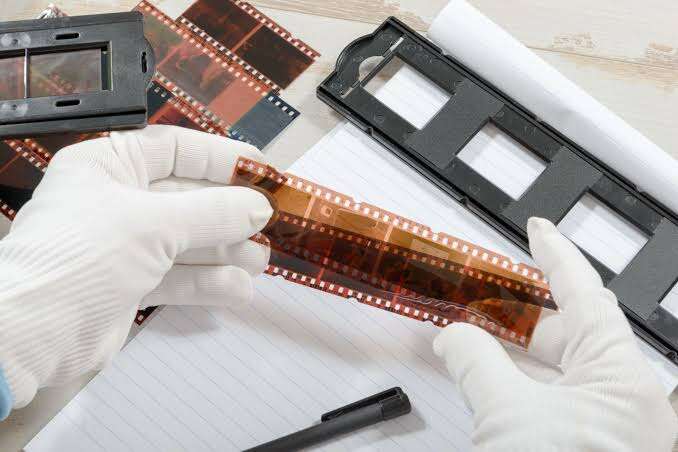 A person wearing white gloves holds photographic film negatives on a desk, surrounded by film holders, a notebook, and a pen.
