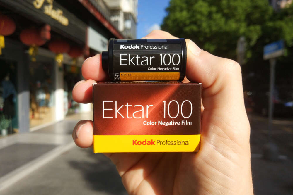 A hand holding a Kodak Professional Ektar 100 color negative film canister and its corresponding box in an urban outdoor setting.