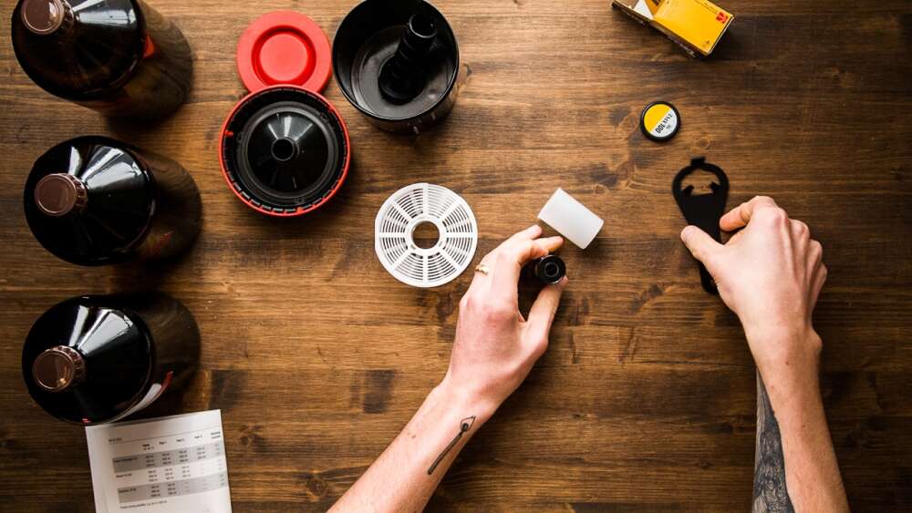 Person's hands working with film developing equipment on a wooden table, surrounded by canisters, film reels, and a developing tank.
