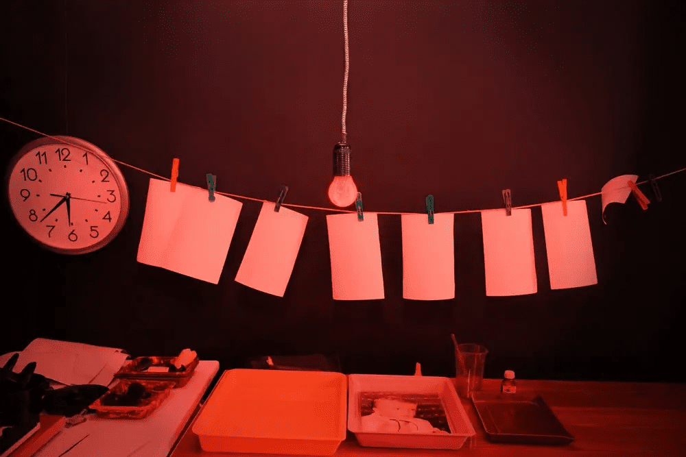 A darkroom setup with a red light illuminating hanging photographic paper on a line, clipped with colourful pegs. Below are trays for developing photos and a clock on the wall showing 7:10.