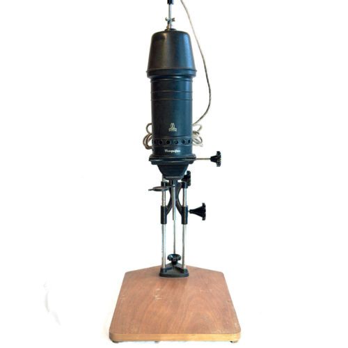 A vintage darkroom enlarger with a black cylindrical body, mounted on a wooden base, featuring adjustment knobs and a power cord.