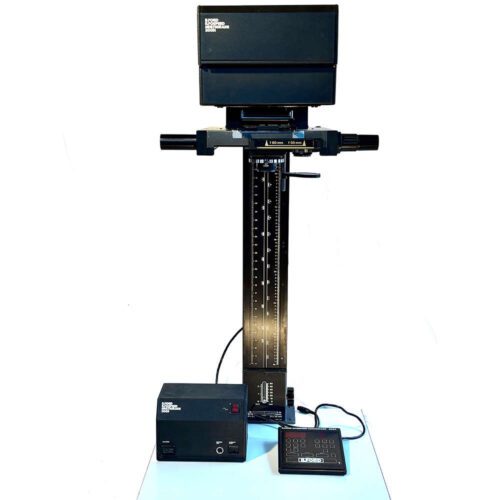 A vintage photographic enlarger with a black column and measuring scale, light housing on top, and control box on the side. The setup is placed on a light background.