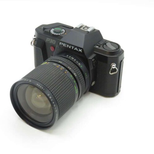 A black Pentax P30 film camera with a large attached zoom lens, angled on a white surface.