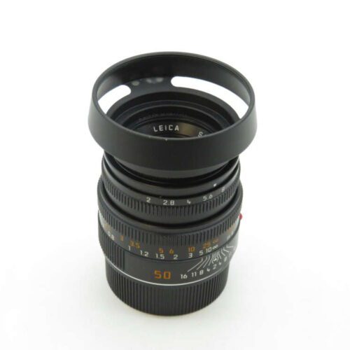 A Leica camera lens with a lens hood attached, showing various aperture and focus markings on the barrel, set against a plain white background.