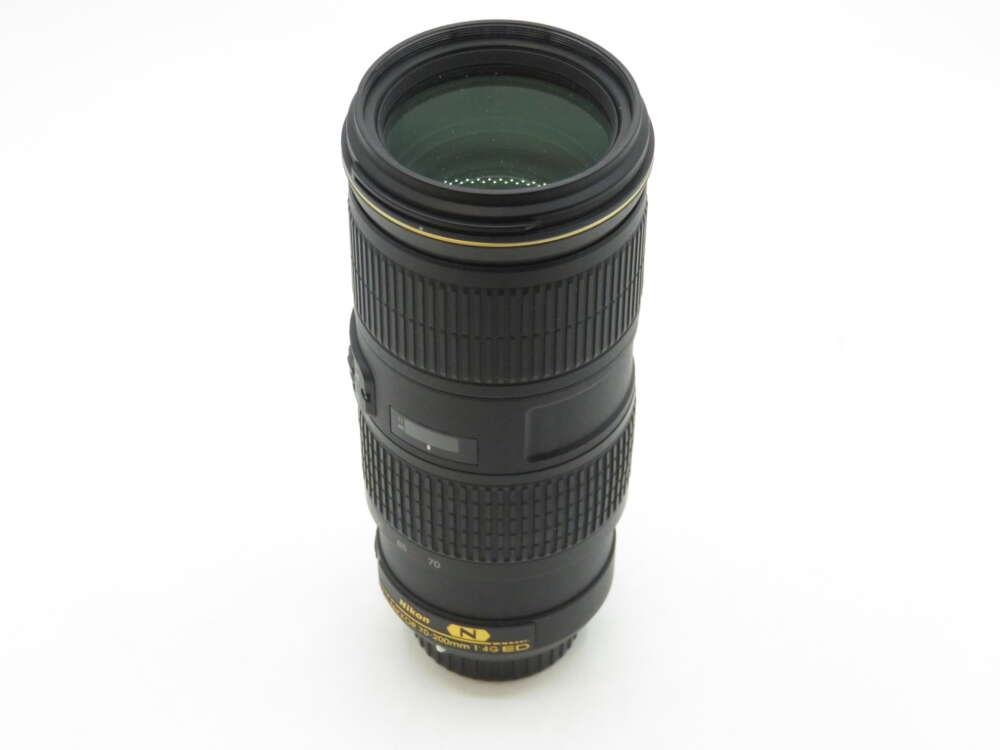 IMG_1343 A black Nikon camera lens with a zoom ring and focusing ring, featuring gold lettering and markings, standing vertically on a plain white background.