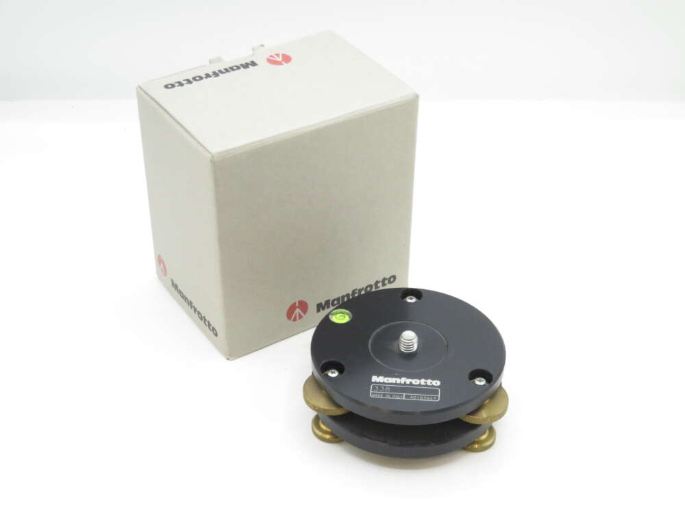 IMG_1387 A Manfrotto levelling base with a threaded screw mount is shown next to its beige box. The base features a black circular design with a built-in spirit level for precision adjustment, suitable for use with tripods or other mounting equipment.