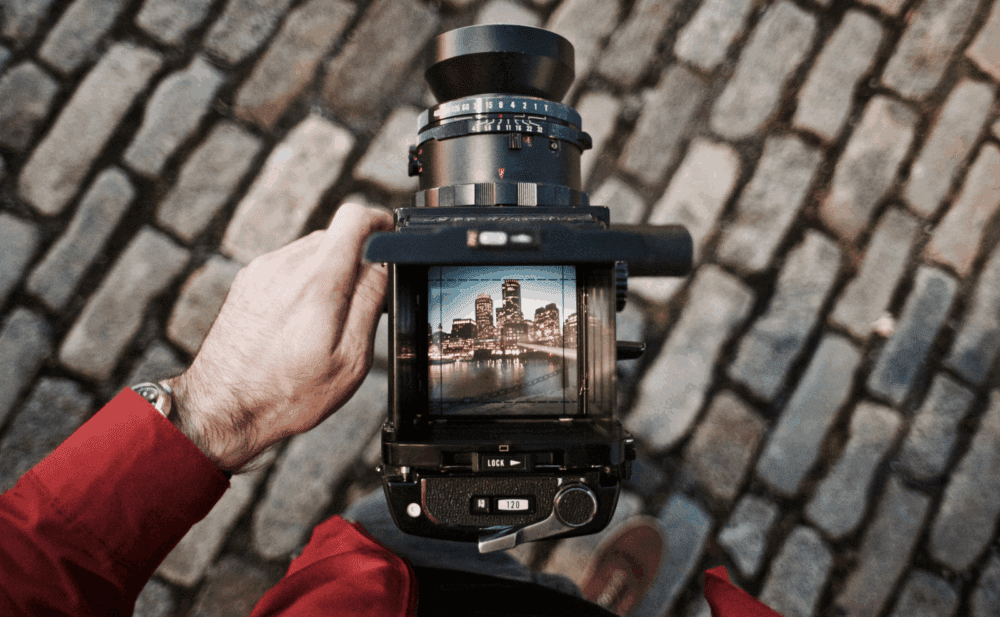 A person in a red jacket is holding a vintage camera, pointing towards cobblestone pavement. The camera's viewfinder displays an image of a city skyline with buildings reflected in water.