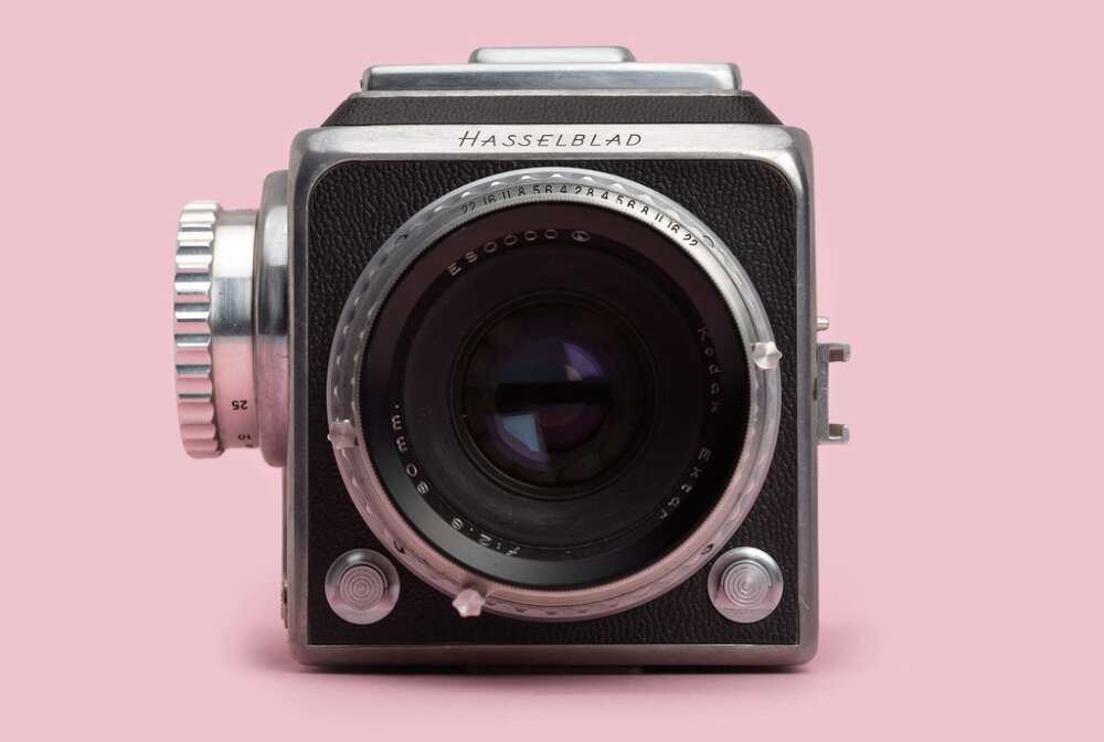 A vintage Hasselblad camera with a black and silver body, displaying a prominent lens in front, set against a pink background.