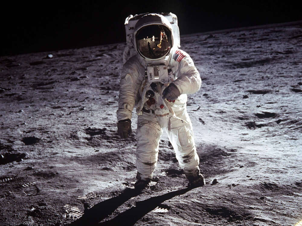An astronaut in a white spacesuit stands on the lunar surface, with the American flag on the arm and a reflection in the helmet visor.