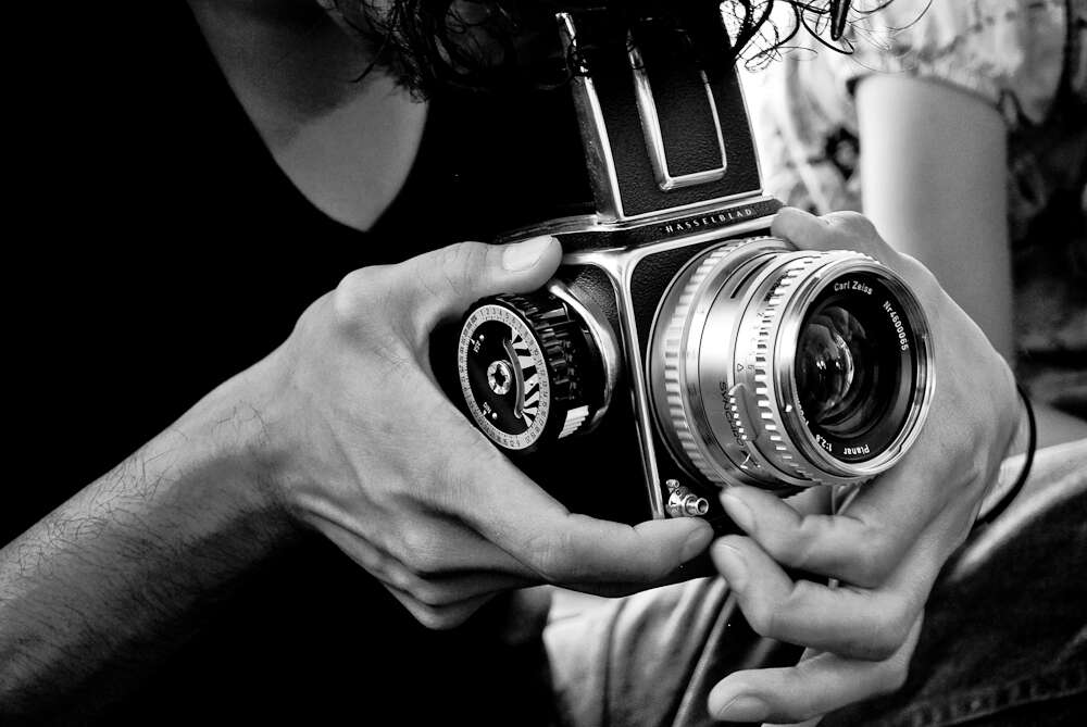 A person holding a vintage Hasselblad camera with a Carl Zeiss lens, captured in black and white, focusing on the detailed camera components.