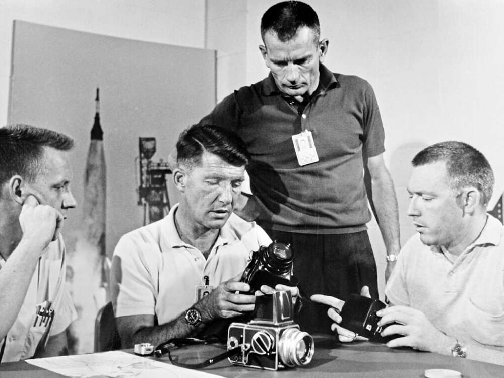 Four men in a room discussing and examining camera equipment, with a poster of a rocket ship in the background.