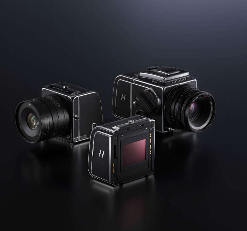 Three Hasselblad medium format camera bodies, two with lenses attached, displayed on a dark reflective surface.