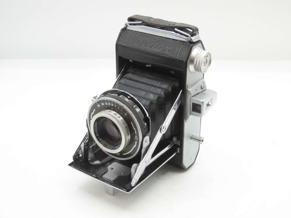 IMG_1492 A vintage folding camera with black leather and silver metal components, featuring a bellows mechanism and a lens with engraved settings and adjustments, displayed against a plain white background.