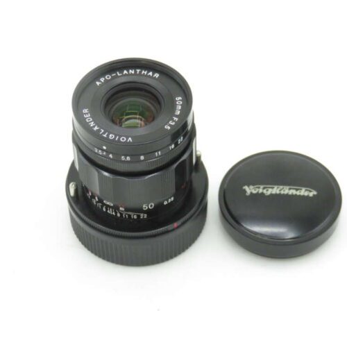 A Voigtländer Apo-Lanthar 50mm f/3.5 camera lens with its detachable lens cap placed beside it, both on a white background.