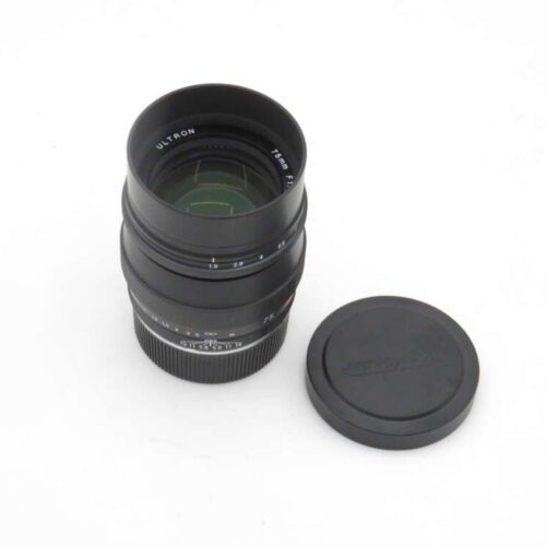A camera lens labeled 'Ultron 75mm F1.8' placed upright next to its black front cap on a white surface.