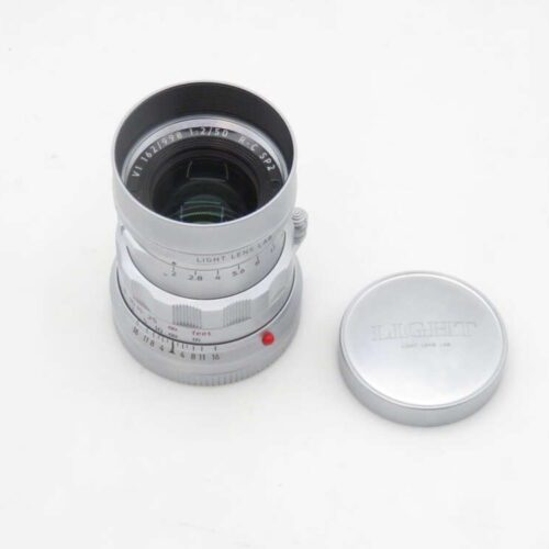 A silver camera lens with marked aperture and focus rings, positioned on a white surface next to its matching lens cap engraved with "LIGHT LENS LAB."