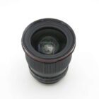 A camera lens with a wide aperture viewed from above, featuring a prominent glass element and a textured focus ring, set against a plain white background.