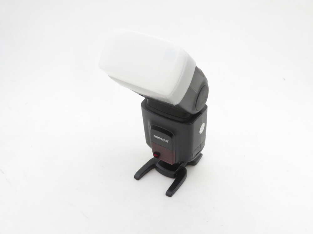 IMG_1638 A camera flash unit with a white diffuser attached, branded "NEEWER," positioned on its stand against a plain white background.