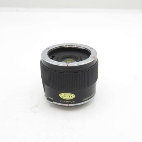 An Olympus teleconverter lens with a black barrel and metallic mount, featuring a JCI certification sticker on its side, placed upright on a plain white surface.