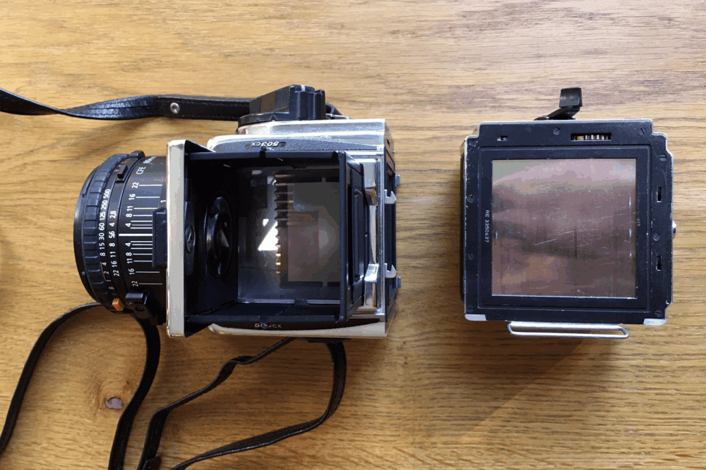 A medium format camera with its back detached, showing the body and lens on the left and the film back on the right, placed on a wooden surface.