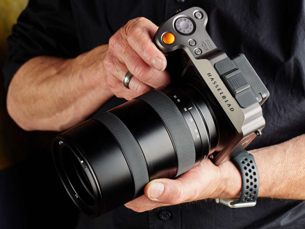 A person holding a Hasselblad camera with a large lens, wearing a ring on their left hand and a smartwatch on their right wrist.