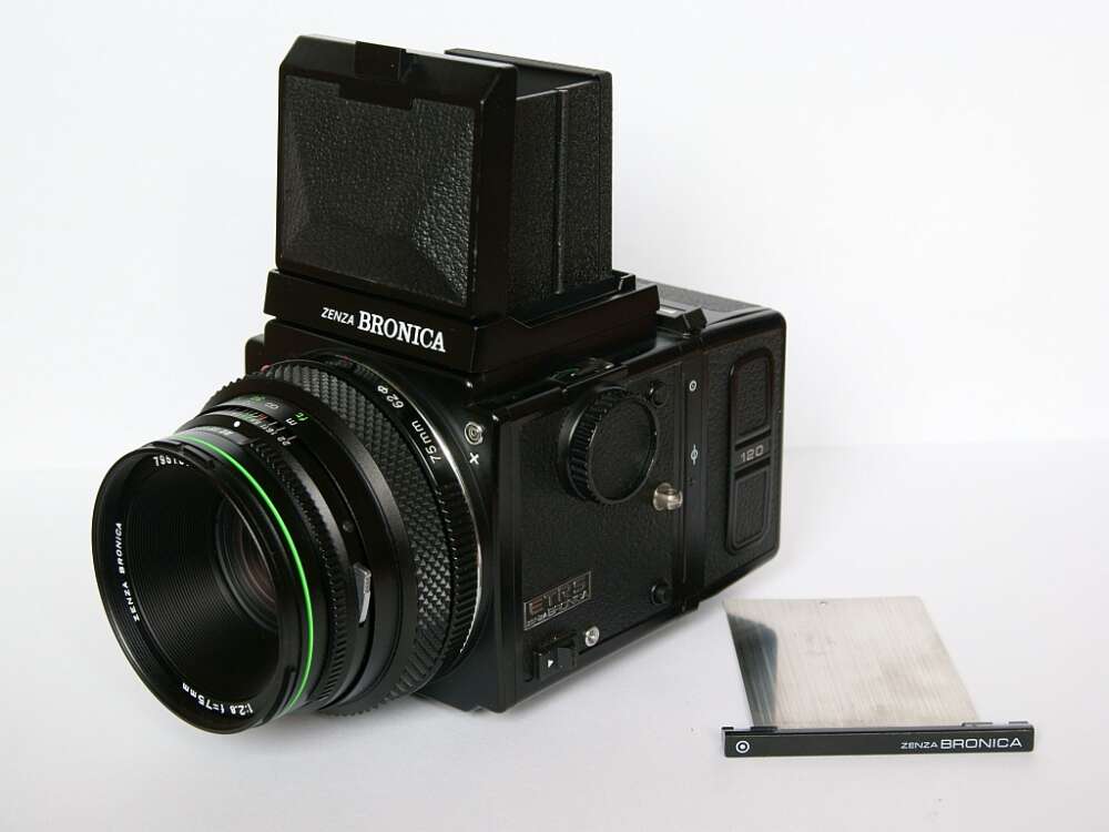 A Zenza Bronica medium format film camera with a detachable viewfinder and a 75mm lens, placed on a white surface. The camera includes a metal dark slide positioned beside it.