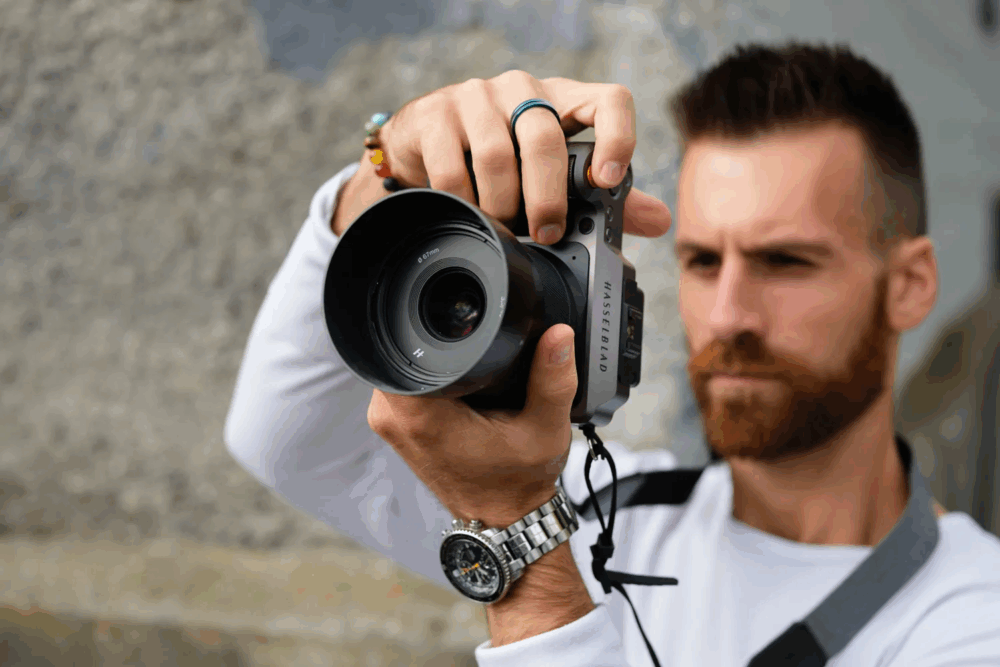 A person wearing a watch and white shirt holds a Hasselblad camera, focusing intently on taking a photo with a blurred background.