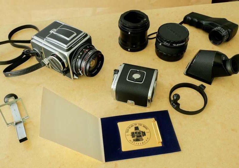 A collection of vintage Hasselblad accessories camera equipment on a beige surface, including the camera body, several lenses, viewfinders, and a gold-plated commemorative plaque.