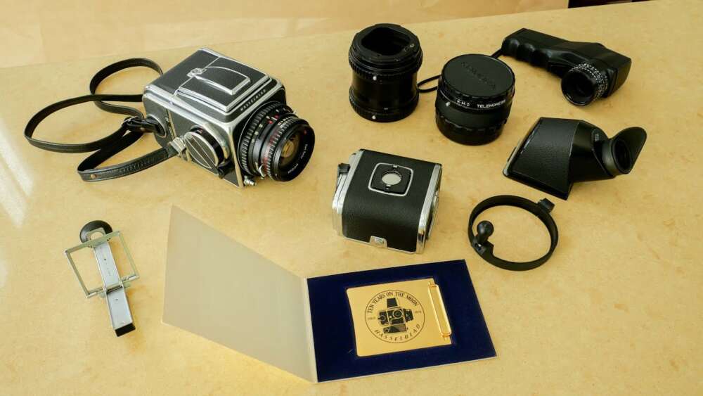 A collection of vintage Hasselblad accessories camera equipment on a beige surface, including the camera body, several lenses, viewfinders, and a gold-plated commemorative plaque.