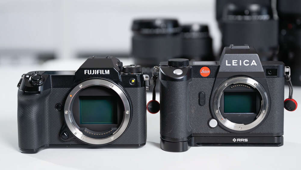 A Fujifilm camera and a Leica camera side by side on a table, both with their lenses removed, revealing the sensor. The cameras are black with textured grips, and are positioned in front of blurred camera lenses in the background.