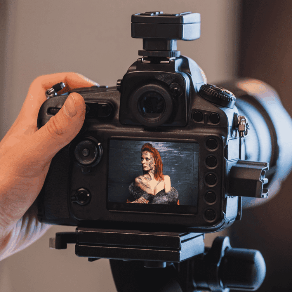 A close-up view of a digital camera's LCD screen displaying a photo of a woman with body paint and feathers, wearing a fur outfit. A hand is holding the camera, which is mounted on a tripod.