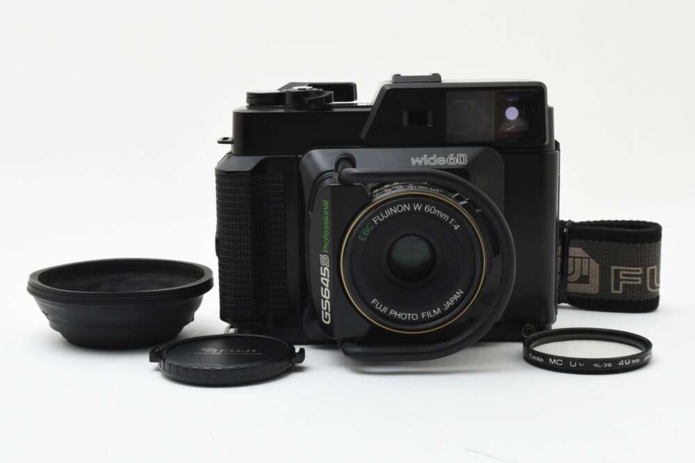 A Fujifilm GS645S Professional medium format film camera with a 60mm f/4 lens, flanked by camera lens caps and filters on a white background.