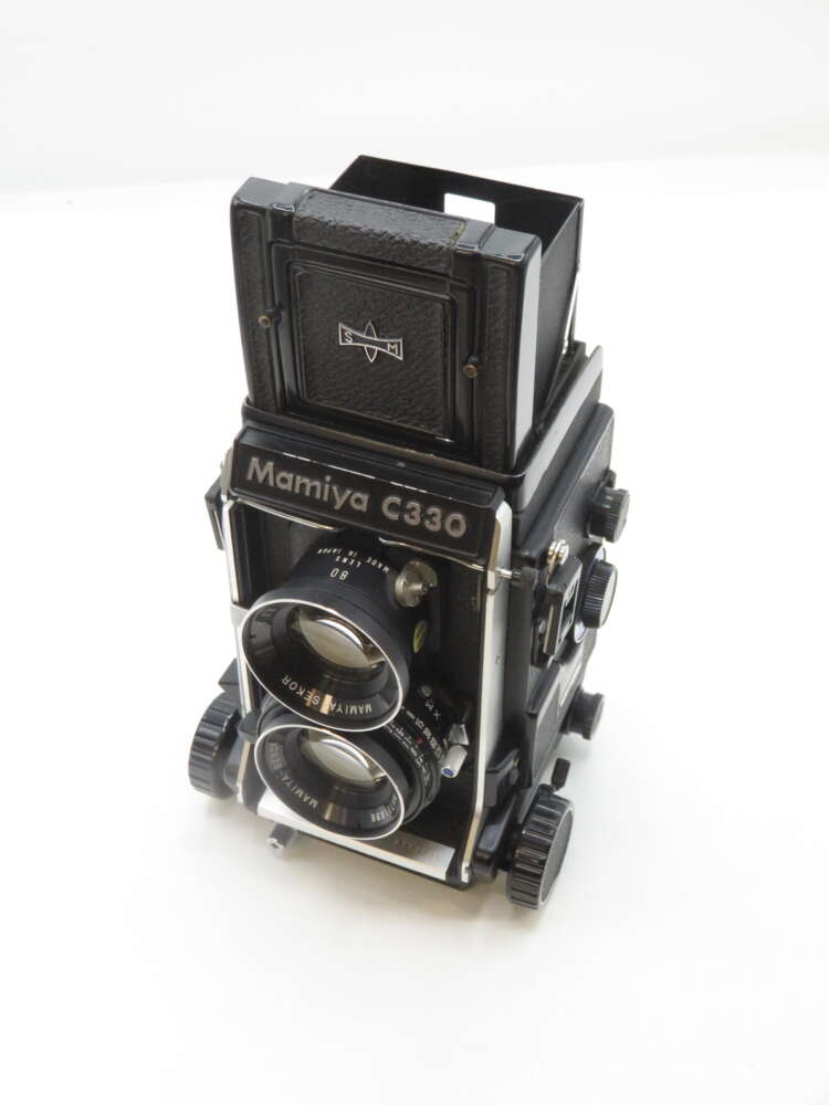 IMG_1774 A vintage Mamiya C330 twin-lens reflex camera with a black body and dual lenses, featuring various knobs and dials for manual adjustments, set on a white background.