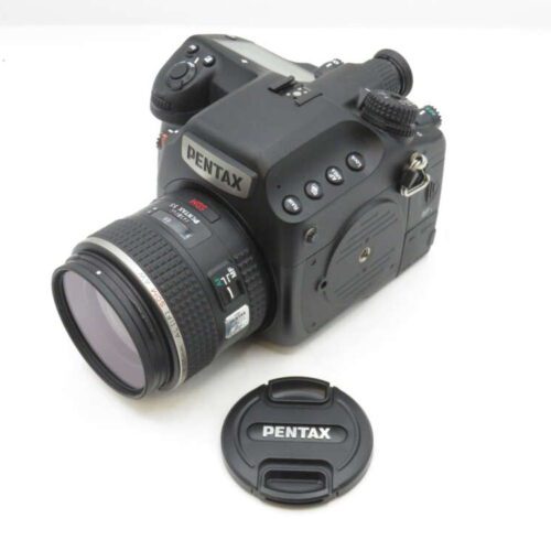 A Pentax camera with a detachable lens is placed on a white surface, with its lens cap lying next to it.