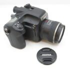 A black Pentax camera with a lens attached, positioned on a white surface. The lens cap, also branded Pentax, is placed nearby.