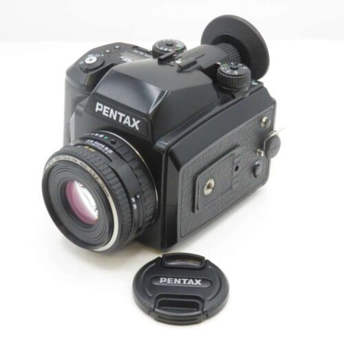 A black Pentax medium format camera with an attached lens, sitting on a white surface. The lens cap is placed in front of the camera.