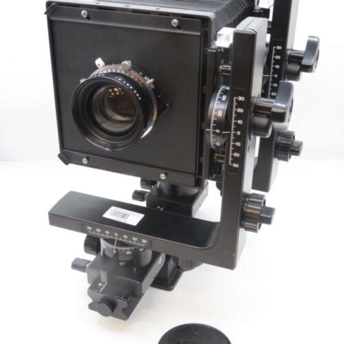 A large format camera with bellows, various adjustment knobs, and a lens at the front. The camera is mounted on a tripod head with precise calibration markings. A lens cap is placed nearby on a white surface.