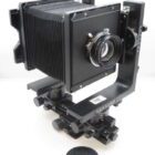 A large format camera with bellows, featuring a black frame, lens, and various adjustment knobs, placed on a white background.