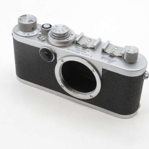 A vintage rangefinder camera body with a black textured exterior and silver metal trim, featuring dials and knobs on the top, with an open lens mount at the front.