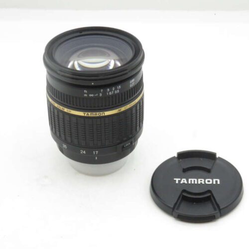 A Tamron zoom lens with a gold band around the middle, next to its lens cap on a white surface.