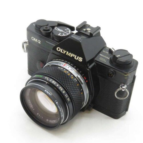 A black Olympus OM-2 film camera with a detachable lens, featuring manual controls and a prominent viewfinder, resting on a plain surface.