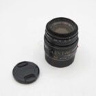 A Leica Summicron-M 50mm f/2 lens with a black body and a detachable lens cap placed beside it, on a plain white background.