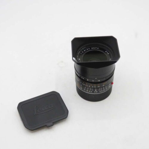 A Leica camera lens with an attached lens hood, placed on a white surface next to a separate Leica lens cap.