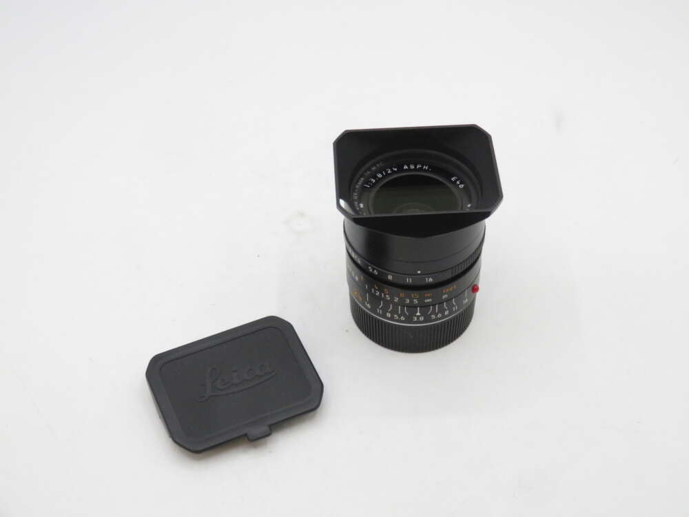 IMG_1966 A Leica camera lens with an attached lens hood, placed on a white surface next to a separate Leica lens cap.