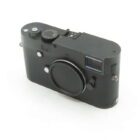 A black rangefinder-style camera with a body cap on the lens mount, featuring a textured grip and various control dials on the top.