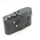 A black rangefinder camera with a minimalist design, featuring a textured grip and a non-removable lens cap. The camera has a top-mounted dial and various buttons on the body for controls, set against a plain white background.