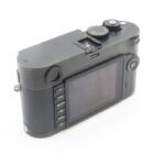 A black Leica digital camera shown from the back, featuring a large LCD screen and several control buttons on the left side of the screen. The top of the camera has a visible dial and a hot shoe mount. The body has a textured grip on one side.
