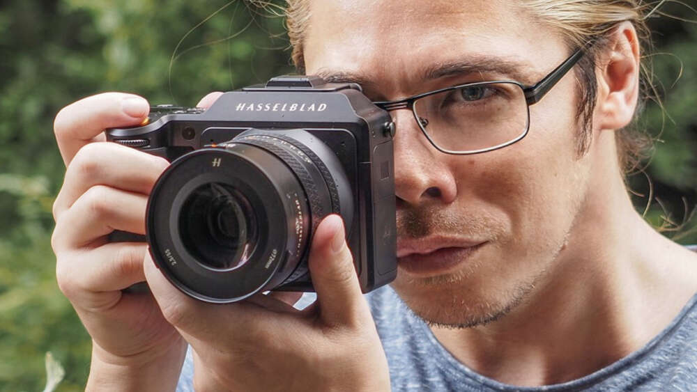 A person holding a Hasselblad camera close to their face, focusing through the viewfinder, with a blurred green background.