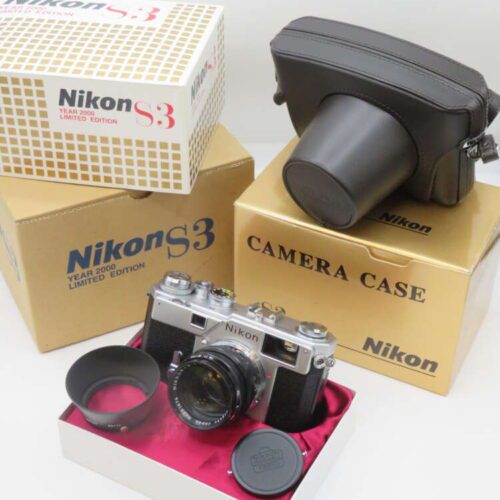 A Nikon S3 Year 2000 Limited Edition camera is displayed alongside its original boxes and a camera case. The camera, featuring a classic silver and black design, is set on a red velvet-lined box with a lens hood and lens cap nearby. Prominent boxes indicate the product as a limited edition item.